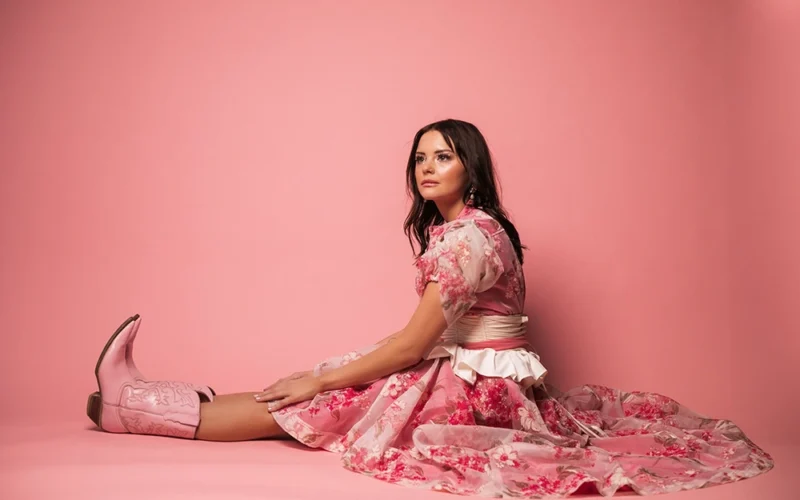 Abbie Callahan seated in a pink dress and boots against a pink background.