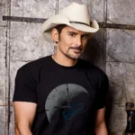 Brad Paisley wearing a cowboy hat and a black T-shirt beside a weathered wall.