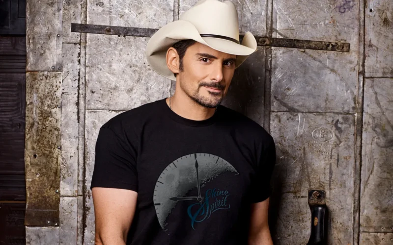 Brad Paisley wearing a cowboy hat and a black T-shirt beside a weathered wall.