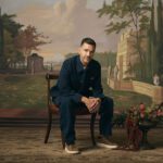 Andy Grammer seated beside a guitar against a painted landscape backdrop