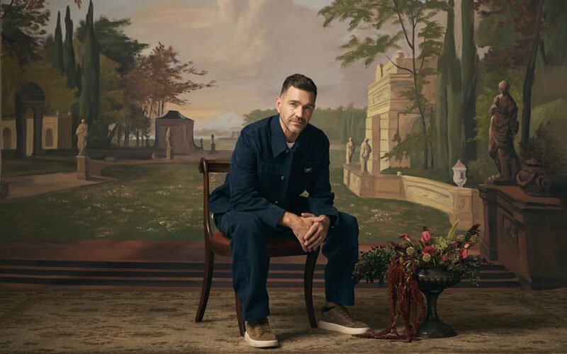Andy Grammer seated beside a guitar against a painted landscape backdrop