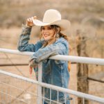 Jenna Paulette wearing a cowboy hat and denim jacket beside a ranch gate
