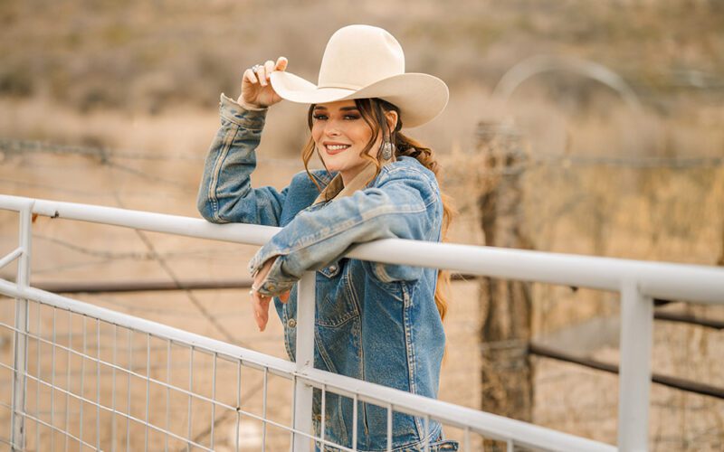 Jenna Paulette wearing a cowboy hat and denim jacket beside a ranch gate