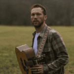 Phillip Phillips carrying a guitar in a grassy field