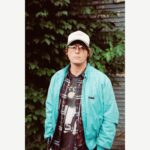 Stephen Wilson Jr. wearing a baseball cap, glasses and a jacket in front of green foliage.
